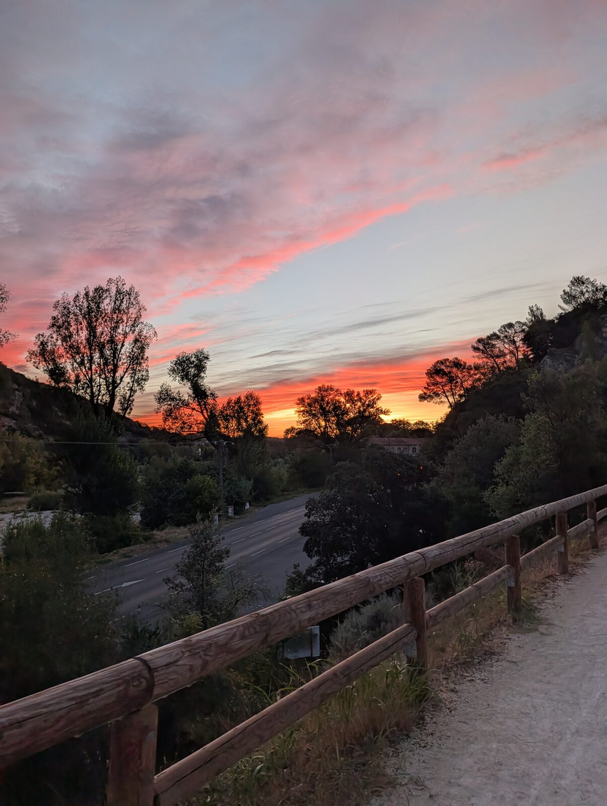 Aube rose et orange au-dessus de la voie verte : barrière en bois au premier plan, route en contrebas, arbres en silhouette sur un ciel strié de nuages roses.
