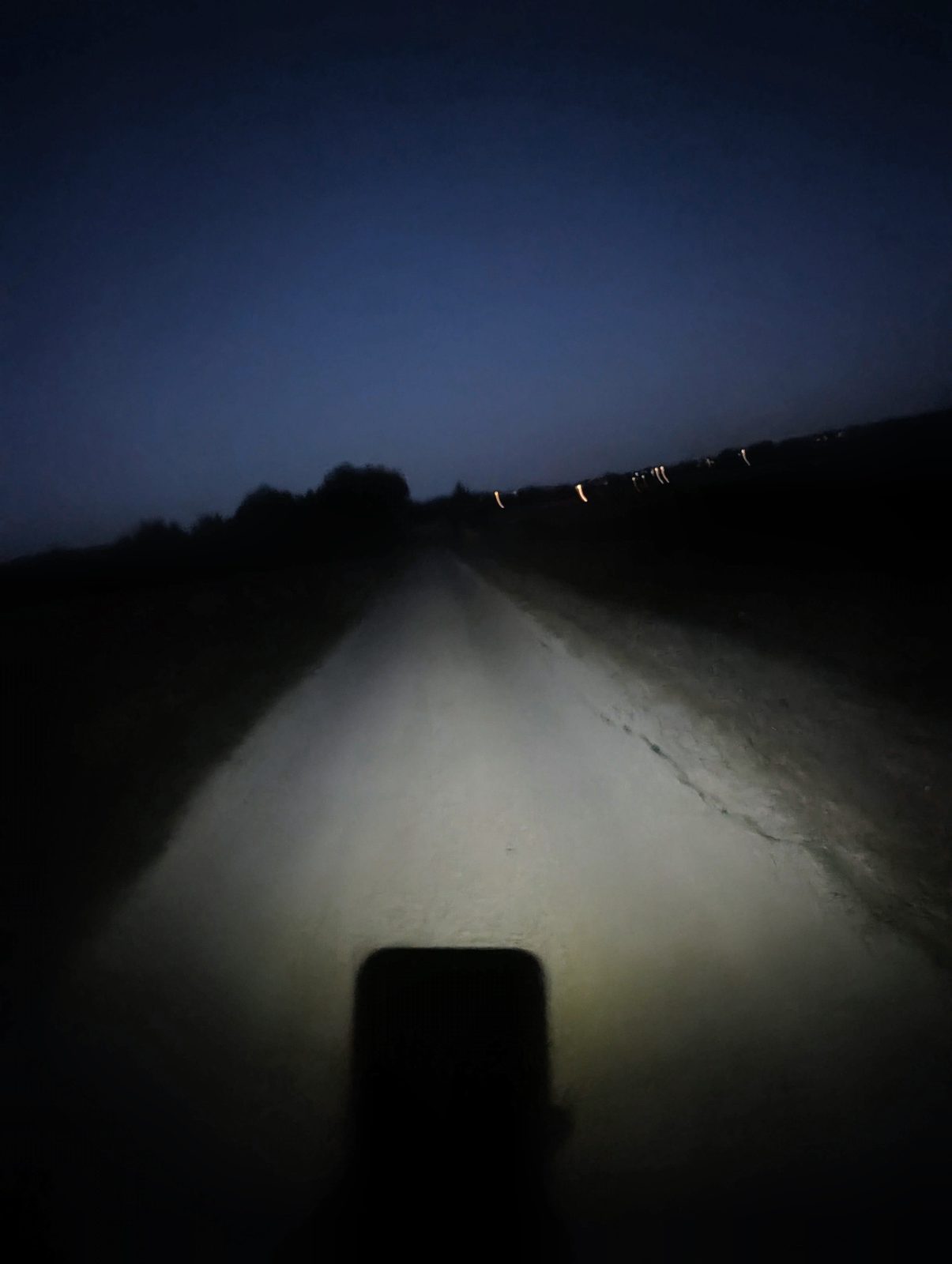 Chemin de terre éclairé par une lampe frontale, dans la nuit, avec les lumières d'un village au loin.