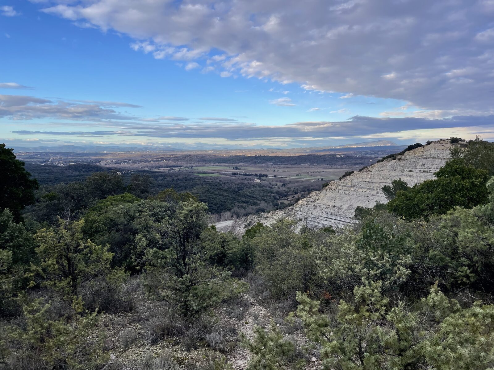 Panorama depuis les hauteurs de garrigue : la vallée du Gard s'étire sous un ciel nuageux, falaise calcaire à droite