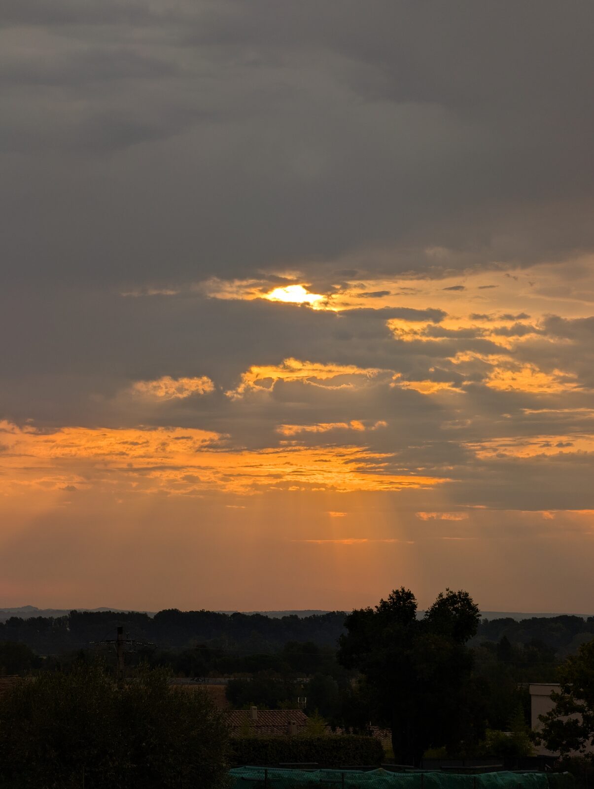 Lever de soleil au départ : le soleil perce entre les nuages gris, des rayons obliques descendent sur l&rsquo;horizon boisé au-dessus des toits.