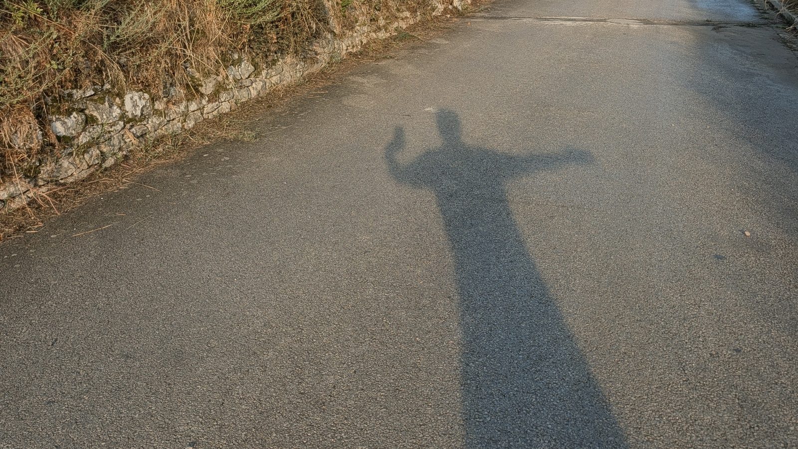 Mon ombre projetée sur la petite route de campagne au soleil rasant du matin, bras levé pour prendre la photo ; muret de pierre et talus d'herbes sèches dorées sur la gauche.
