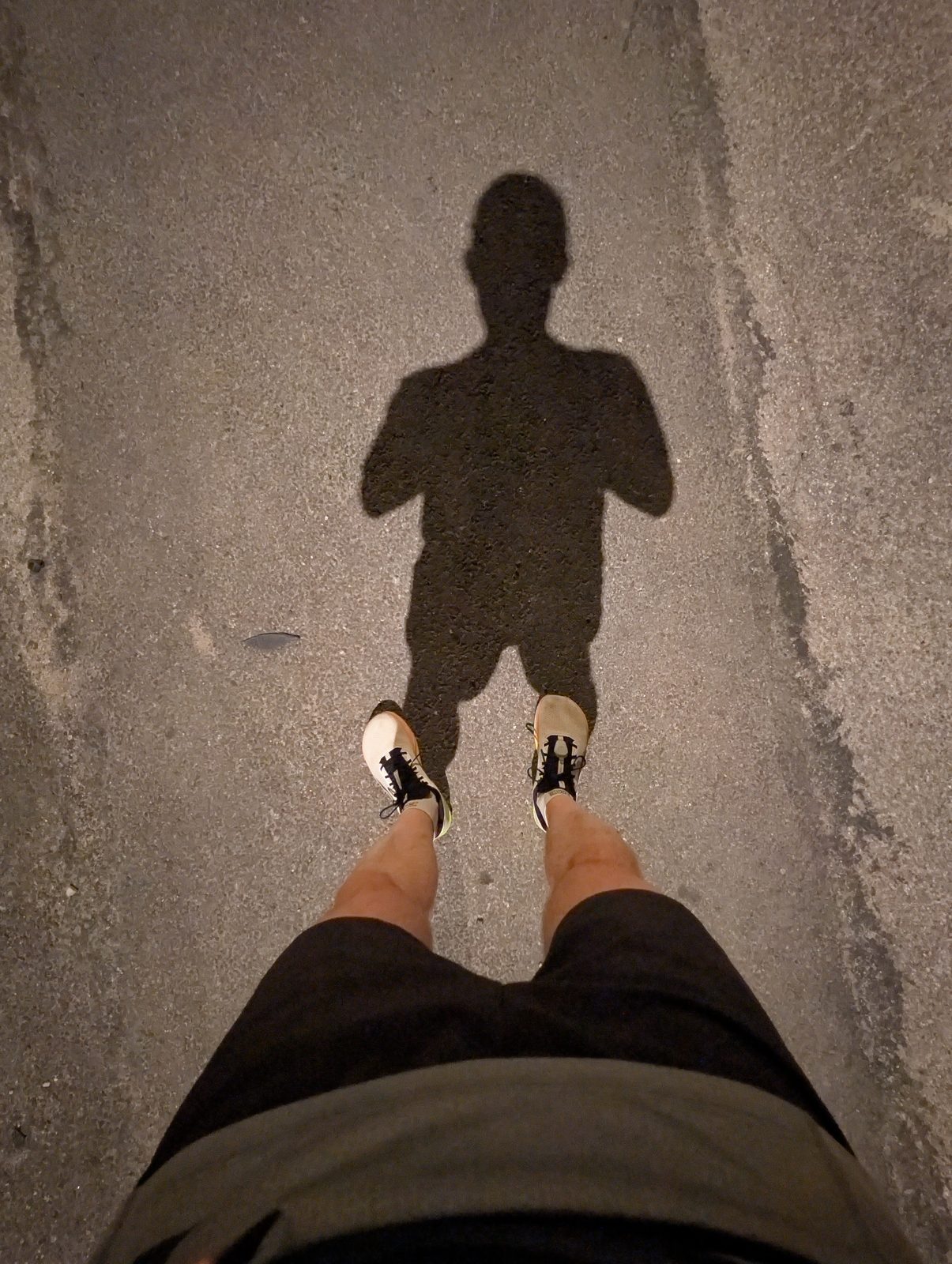 Vue plongeante sur mes chaussures minimalistes blanches et mon ombre projetée sur le bitume par un lampadaire, avant le lever du jour.
