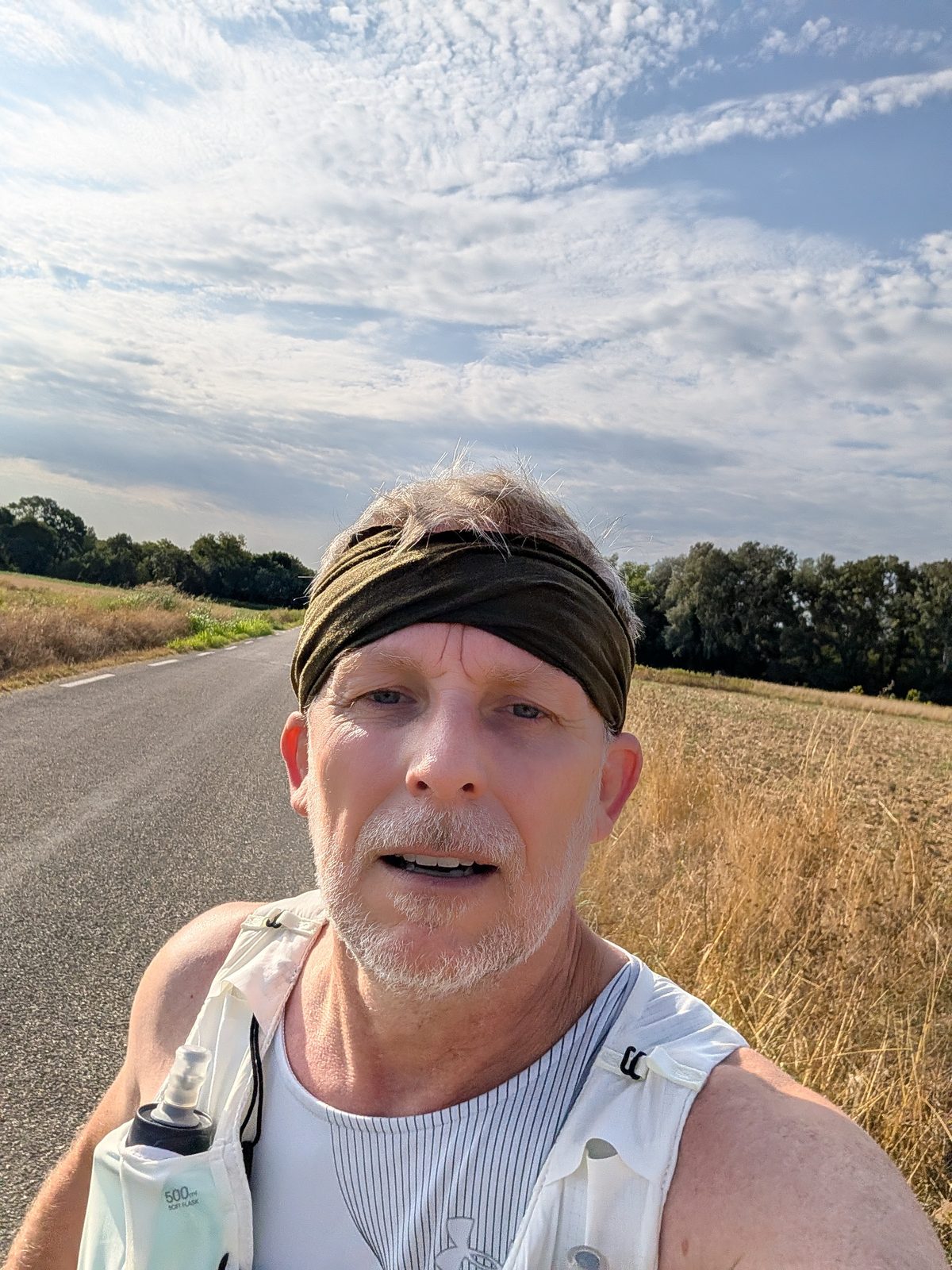 Selfie en pleine sortie sur une petite route de campagne, gilet d'hydratation blanc et soft flask de 500 mL sur la poitrine, bandeau kaki, champs de chaumes dorés et ciel voilé de fin de matinée.