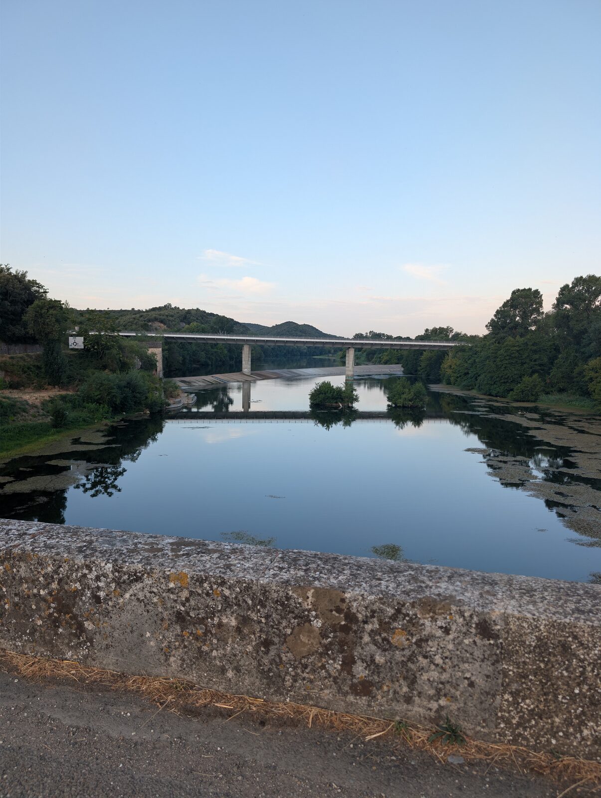 La rivière au petit matin, lisse comme un miroir, un pont routier qui la traverse au loin et se reflète dans l&rsquo;eau.