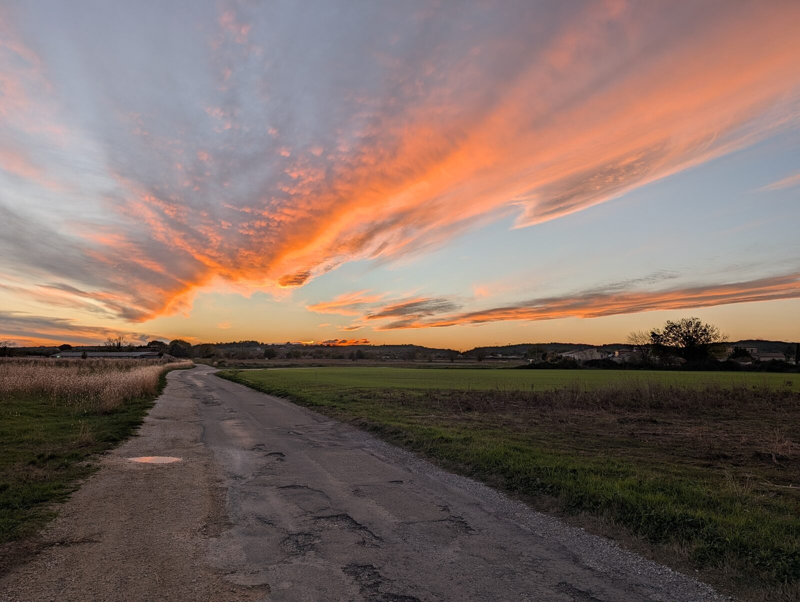 Une petite route de campagne file vers l&rsquo;horizon sous un ciel de coucher de soleil embrasé d&rsquo;orange.