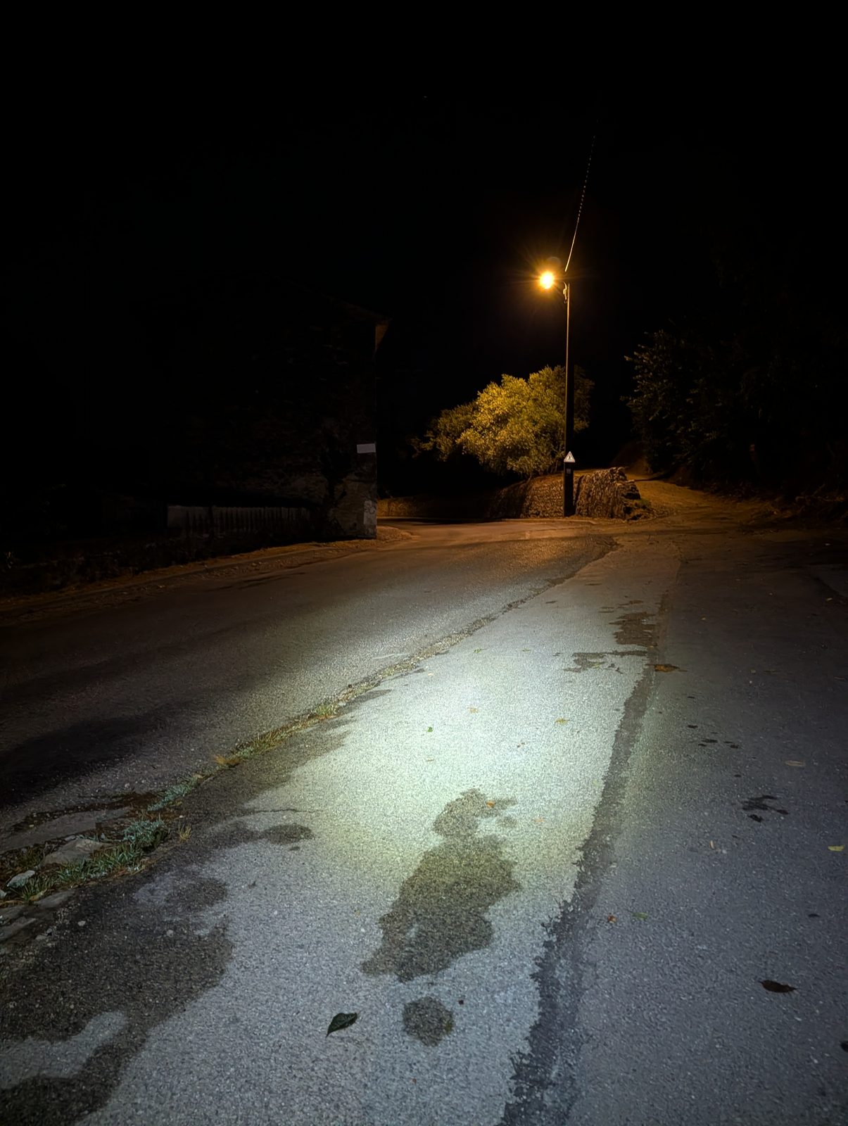 La petite route du départ en pleine nuit, éclairée par un lampadaire orange ; l'asphalte luit dans le faisceau, un arbre et un muret de pierre sortent de l'obscurité.