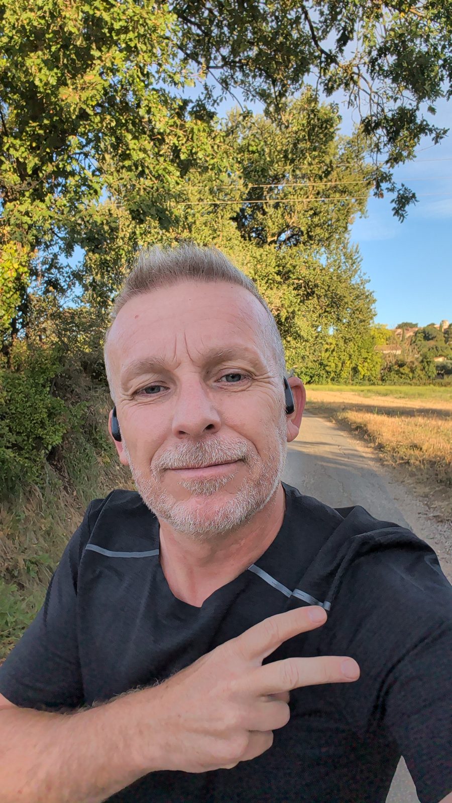 Selfie en courant sur un chemin entre les arbres et un champ, tee-shirt noir, écouteurs à conduction osseuse, soleil rasant du matin.