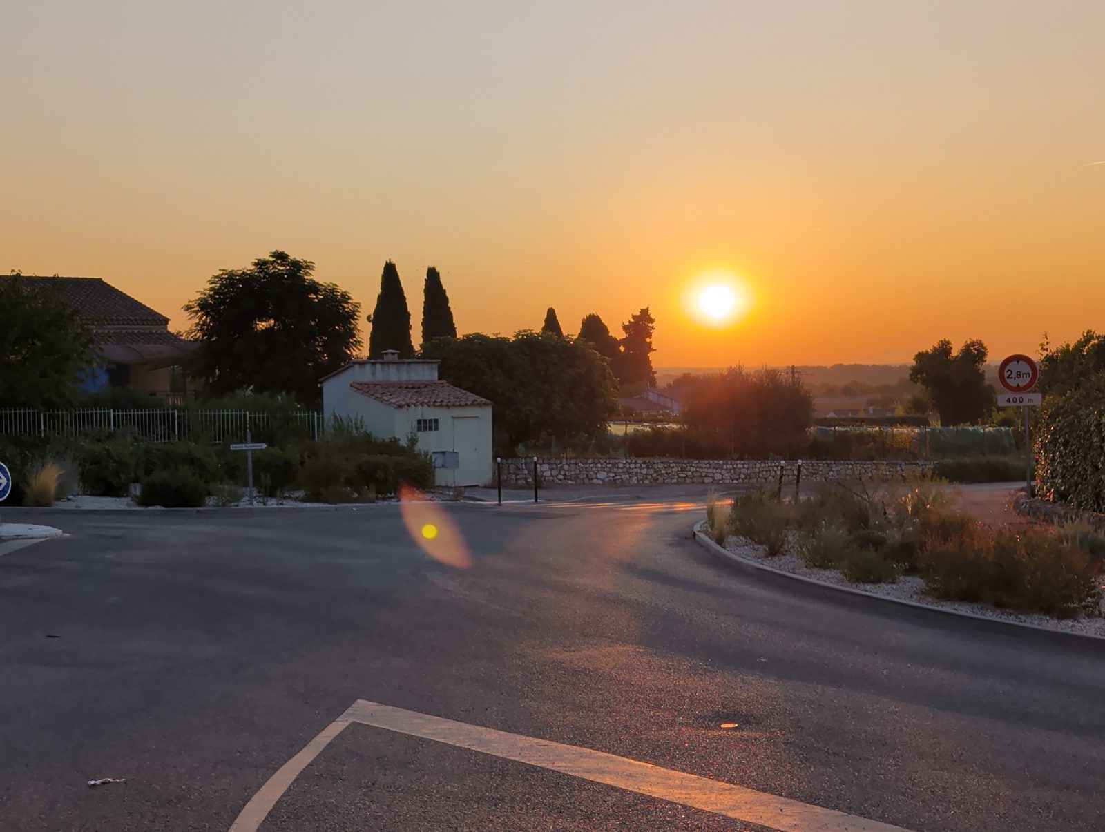Lever de soleil orangé sur une route de village du Gard, cyprès en silhouette et mur de pierres sèches, tôt le matin.